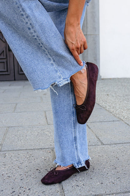Person in frayed light blue jeans adjusts MIA Tinna Bow Ballet Flats in brown while standing on a stone sidewalk.