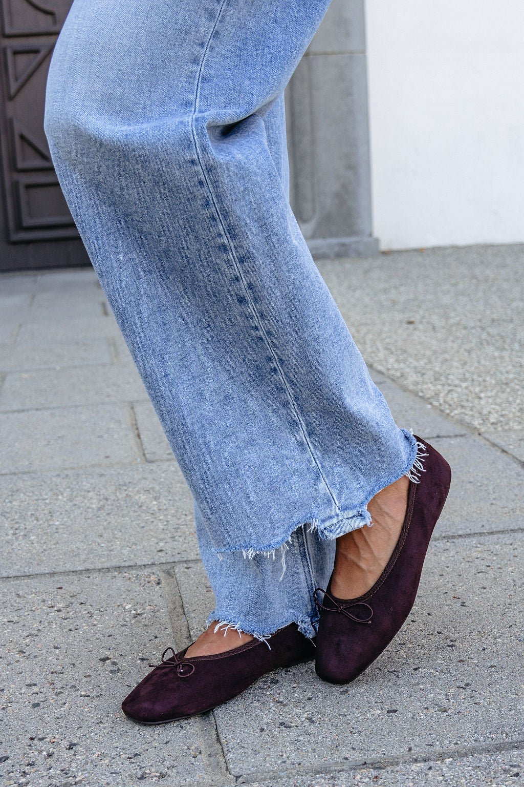 Individual in frayed hem jeans and MIA Tinna Bow Ballet Flats in brown, standing on a stone pavement.
