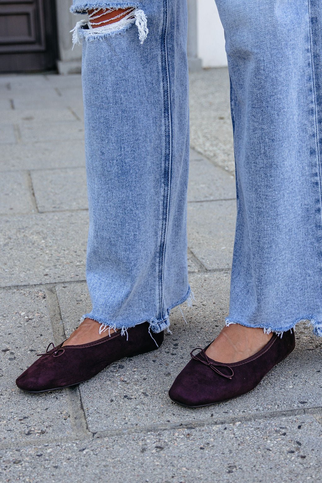 Person in ripped, light blue jeans and MIA Tinna Bow Ballet Flats - Brown stands on a concrete sidewalk.