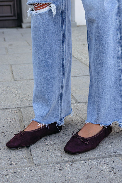 Person in ripped, light blue jeans and MIA Tinna Bow Ballet Flats - Brown stands on a concrete sidewalk.