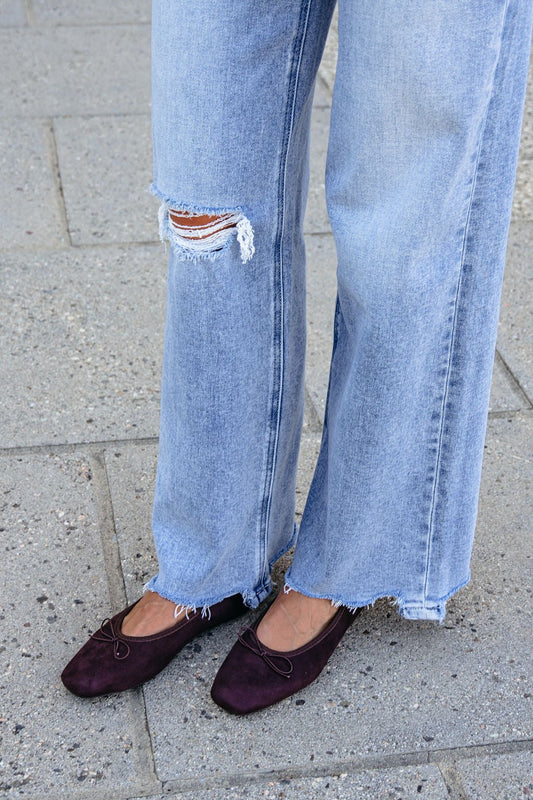 Person in ripped light blue jeans and MIA Tinna Bow Ballet Flats - Brown standing on a paved sidewalk.