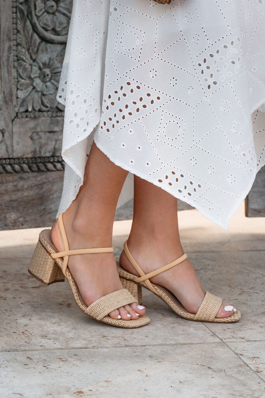 Woman in a white eyelet dress wears MIA Zissy Natural Strappy Heels, standing on a stone floor for a chic block heel look.