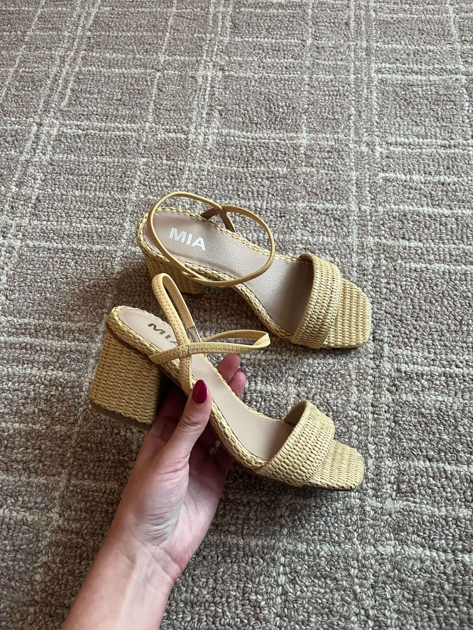 A hand holds MIA Zissy Natural Strappy Heels with woven block heels on a textured beige carpet.