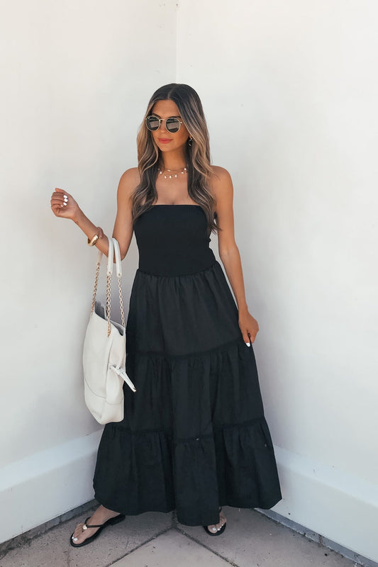 Woman in a Midnight Ease Strapless Tiered Maxi Dress - Black, with sunglasses, black sandals, and white bag, by a white wall.