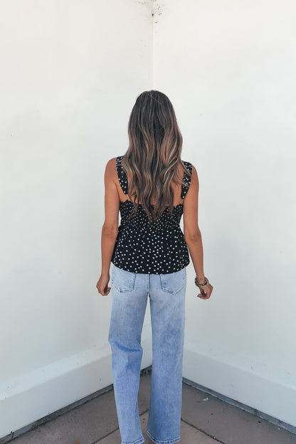 Woman with long wavy hair in a Midnight Lace Print Tank Top - Black and light blue jeans stands facing a white corner wall.