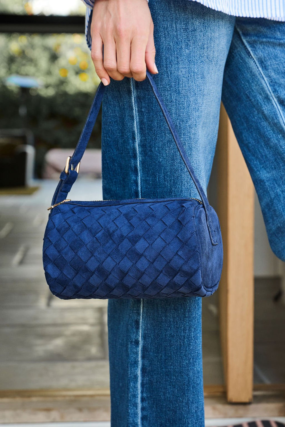 A person in blue jeans holds the Midnight Textured Vegan Leather Shoulder Bag by its strap while standing near an open door.