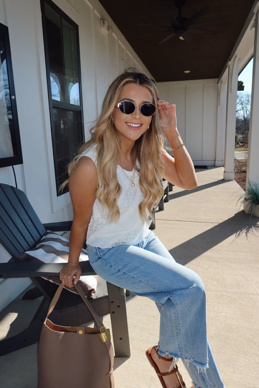 A woman with long blonde hair in sunglasses and a Mika Ivory Ruffled Lace Top sits on a porch chair, smiling with a brown handbag.