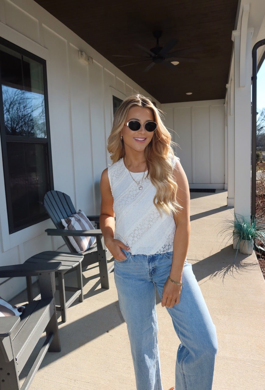 Woman with long blonde hair in sunglasses wears the Mika Ivory Ruffled Lace Top and blue jeans, standing on a sunny porch.
