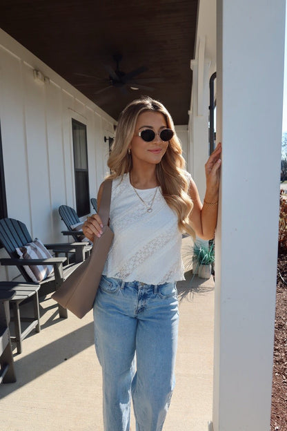 A woman in sunglasses, relaxed jeans, and the Mika Ivory Ruffled Lace Top stands on a porch holding a taupe bag by a white column.