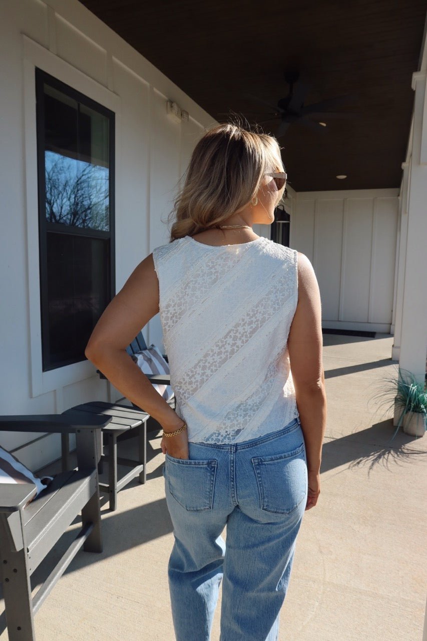 A woman in the Mika Ivory Ruffled Lace Top and blue jeans stands on a sunny porch, facing away.
