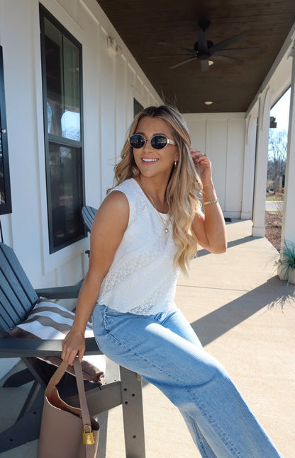 Smiling woman in sunglasses wears the Mika Ivory Ruffled Lace Top, sitting on a porch chair holding a brown bag.