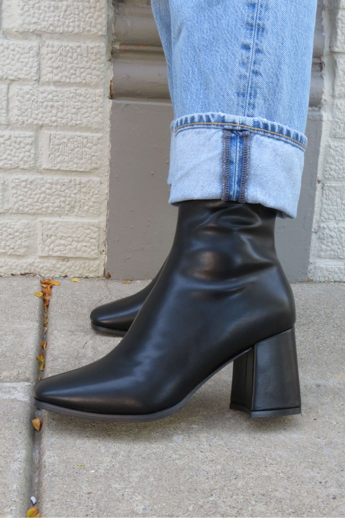 Milan Black Chunky Ankle Booties styled with light blue cuffed jeans, standing on a sidewalk by a textured wall.