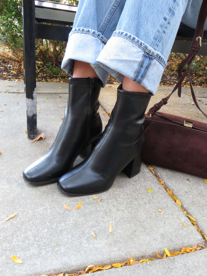 Person wearing cuffed blue jeans and Milan Black Chunky Ankle Booties sits on a bench by a brown handbag among autumn leaves.