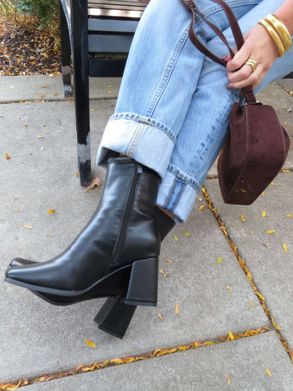 A person in cuffed blue jeans and gold bangles sits on a sidewalk, holding a brown handbag and wearing Milan Black Chunky Ankle Booties.