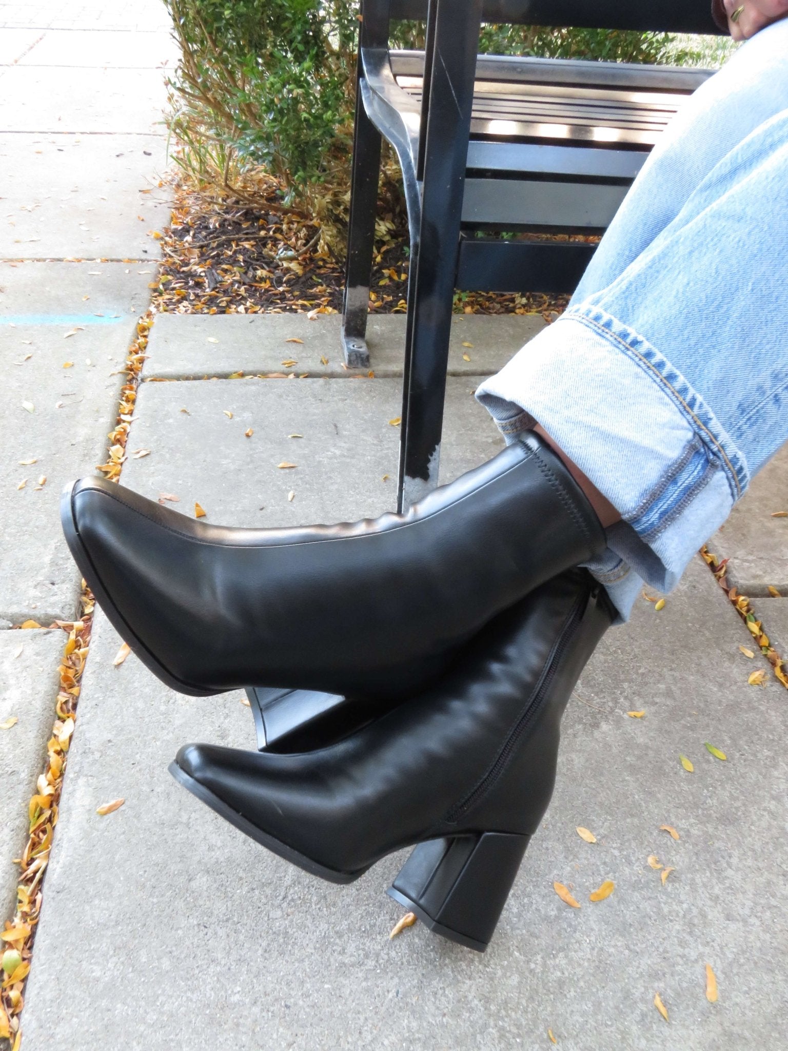 Person wearing Milan Black Chunky Ankle Booties and light blue jeans, sitting cross-legged on a leaf-strewn sidewalk bench.