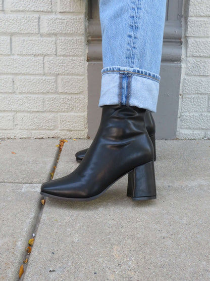 Wearing light blue cuffed jeans, a person stands by a white brick wall in Milan Black Chunky Ankle Booties.