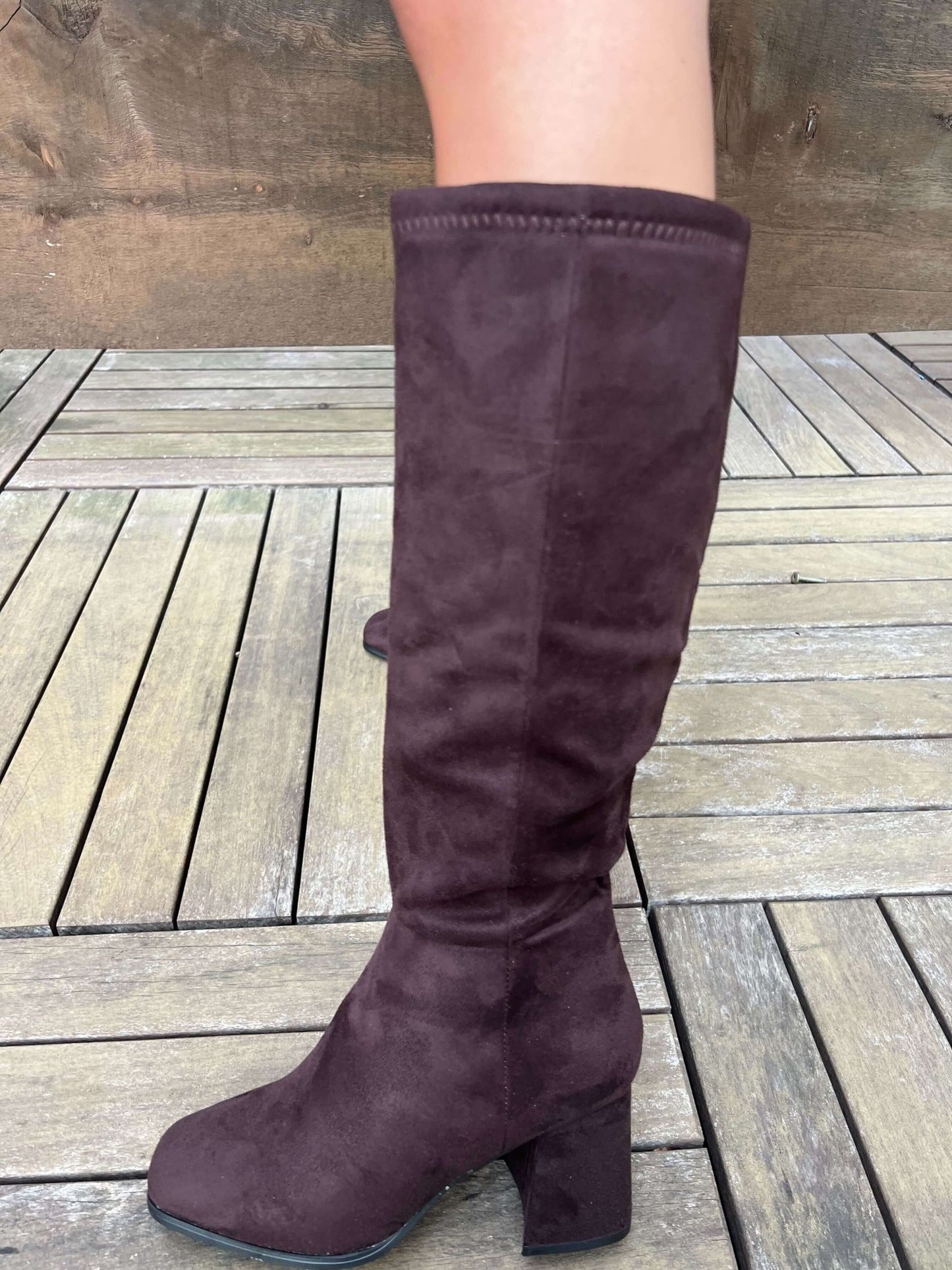 Someone in Milan Brown Heeled Knee High Boots stands on a wooden deck, wearing dark brown faux suede with a block heel.