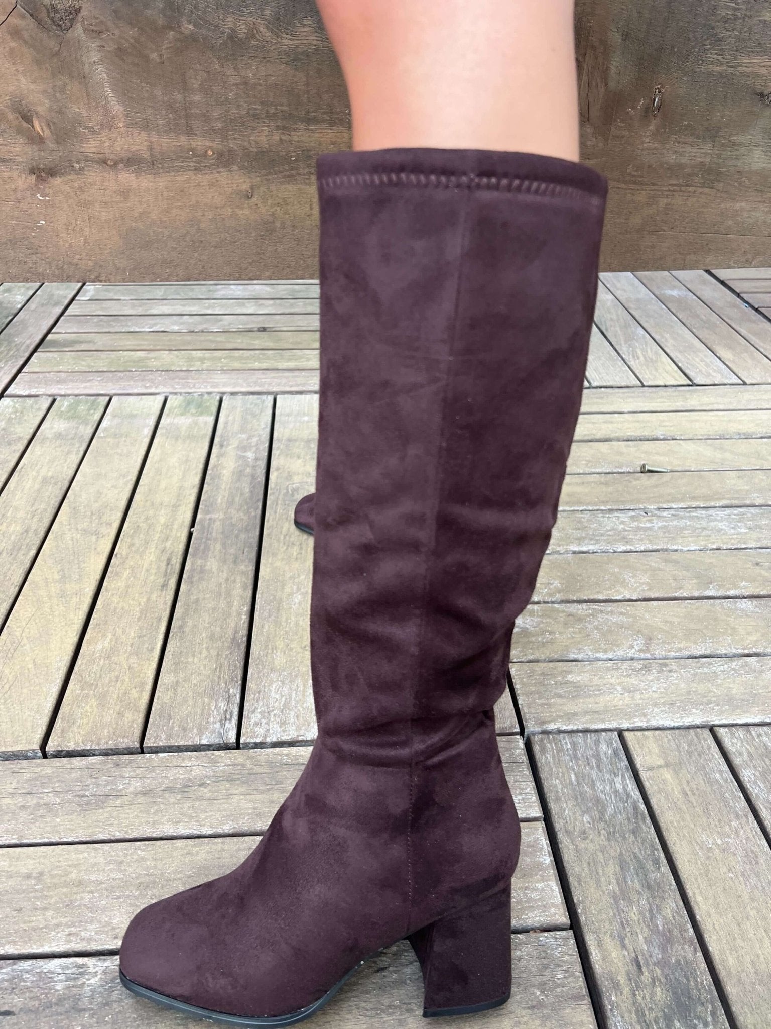 Someone in Milan Brown Heeled Knee High Boots stands on a wooden deck, wearing dark brown faux suede with a block heel.