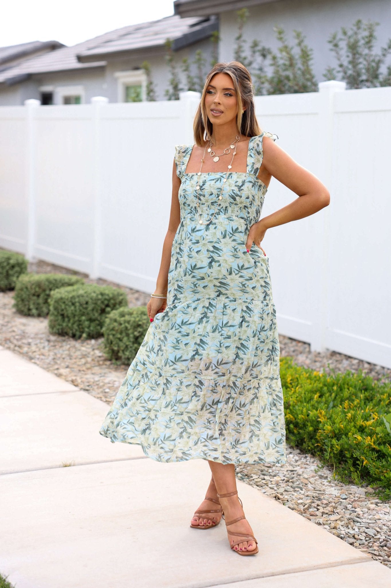 Woman smiles at the camera in a Mint Green Floral Print Smocked Midi Dress, standing by a white fence and greenery.
