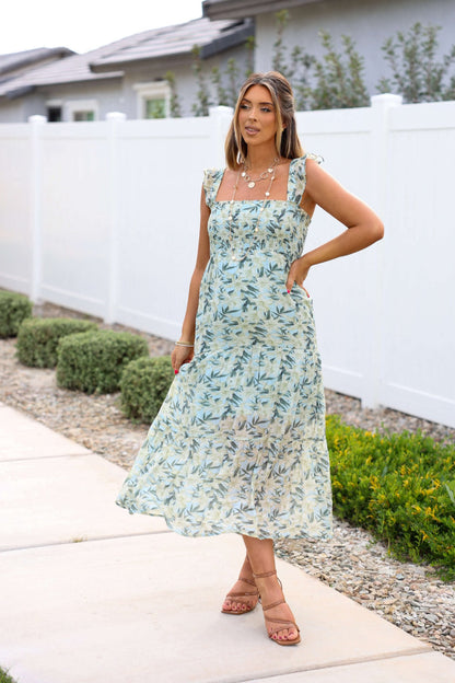 Woman smiles at the camera in a Mint Green Floral Print Smocked Midi Dress, standing by a white fence and greenery.