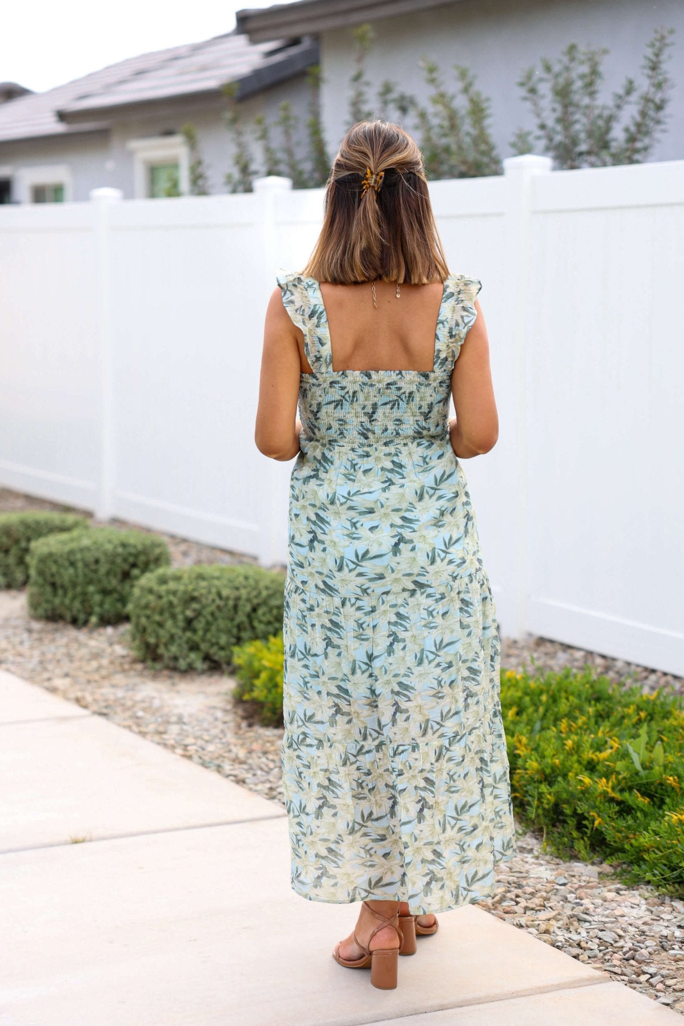 A woman in a Mint Green Floral Print Smocked Midi Dress and sandals walks along a path by a white fence and green shrubs.