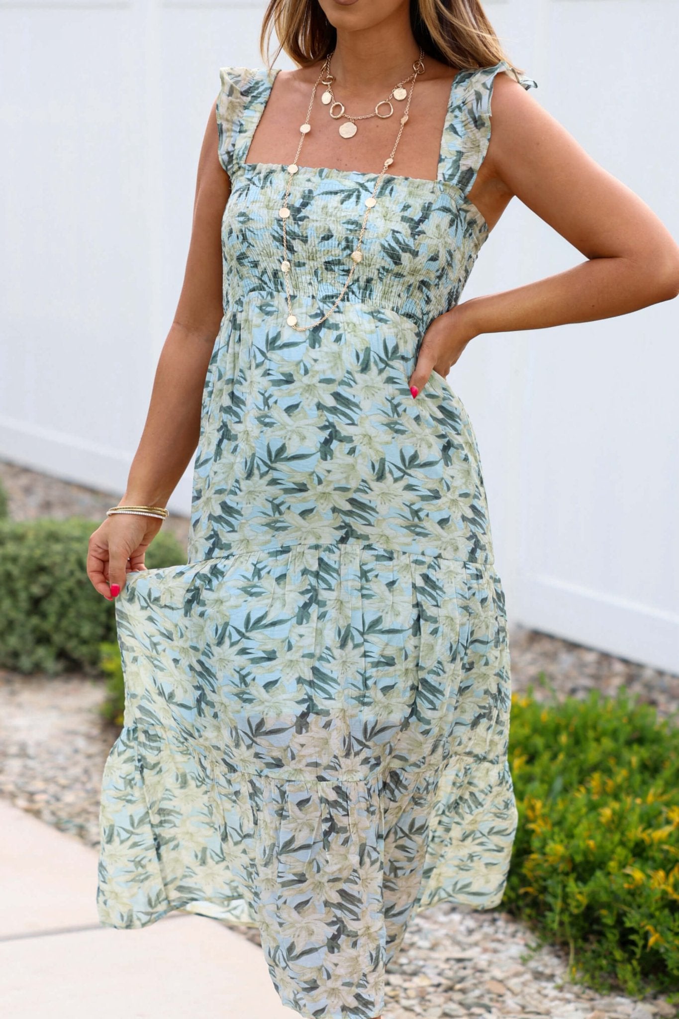 Woman in a bump-friendly Mint Green Floral Print Smocked Midi Dress, layered gold necklaces, outdoors by greenery and a white fence.