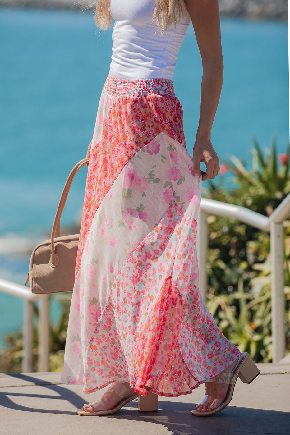 Woman wears a white tank top and the Mixed Print Smocked Maxi Skirt in pink near a seaside railing, holding a beige bag.