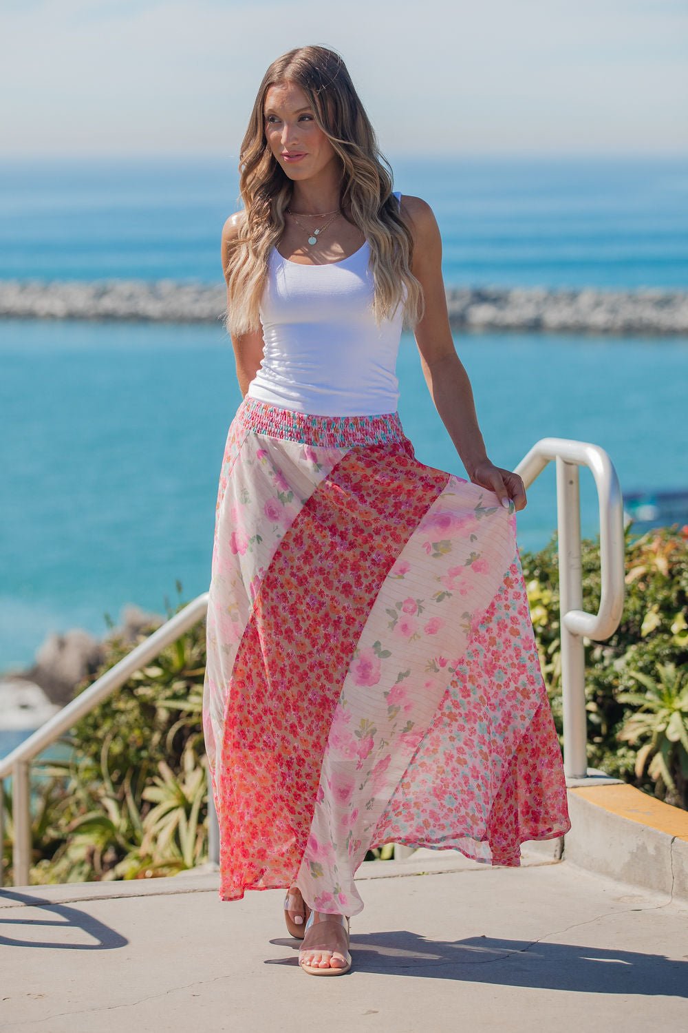 A woman wears the Mixed Print Smocked Maxi Skirt in pink outdoors by the ocean on a sunny day.