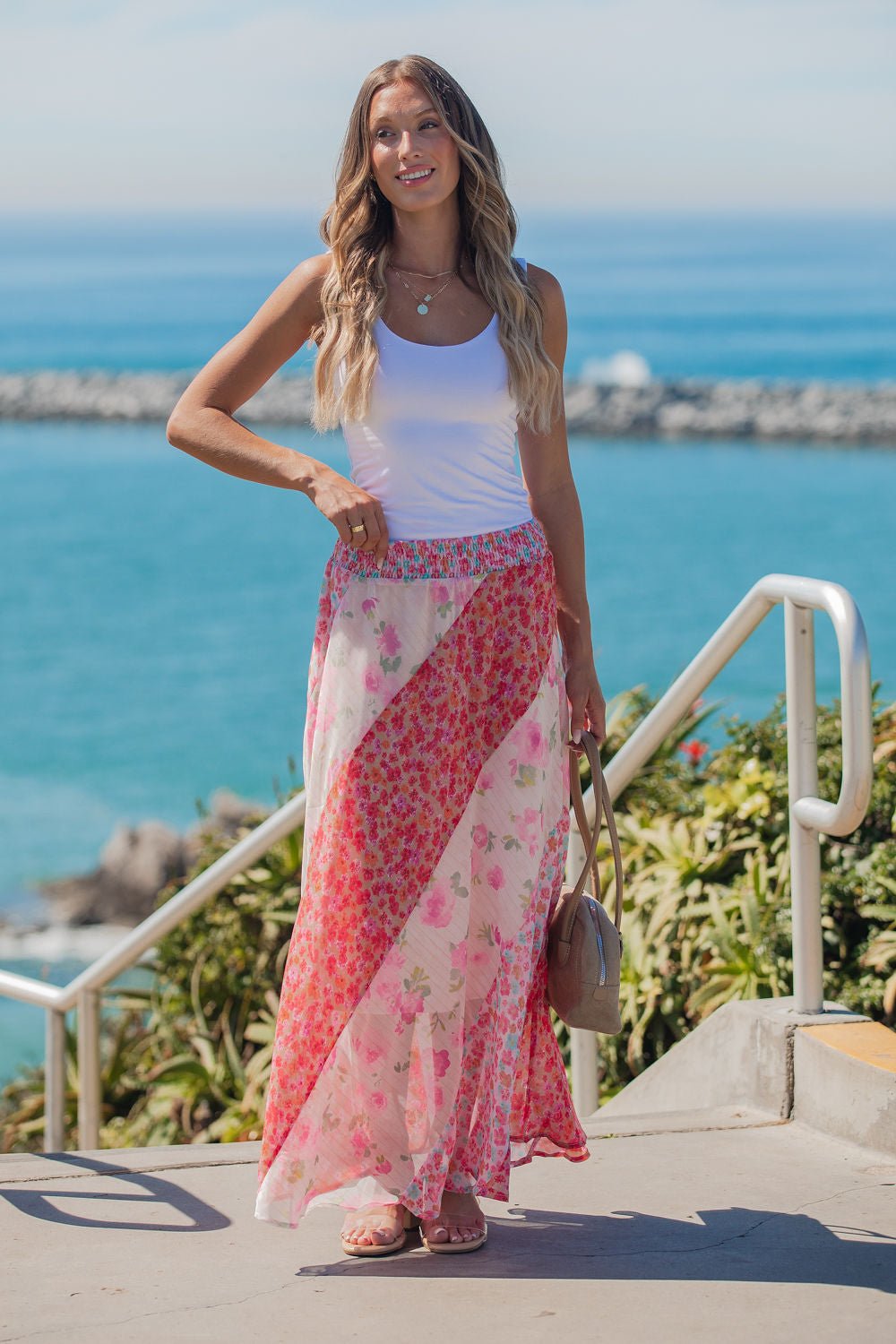 Woman in a white tank and Mixed Print Smocked Maxi Skirt - Pink stands outdoors by the ocean, smiling on a sunny day.