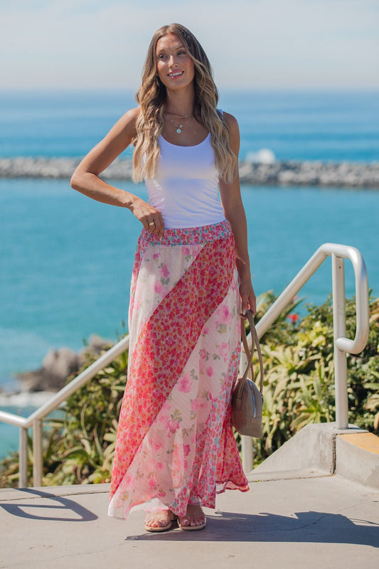 A woman in a white tank top and Mixed Print Smocked Maxi Skirt - Pink stands outdoors by the ocean, smiling with a handbag.