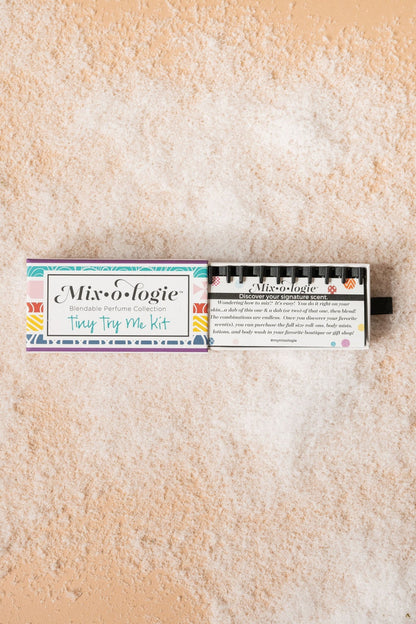 The Mixologie Tiny Try Me Perfume Kit rests on sand, offering a customizable fragrance experience in a compact set.