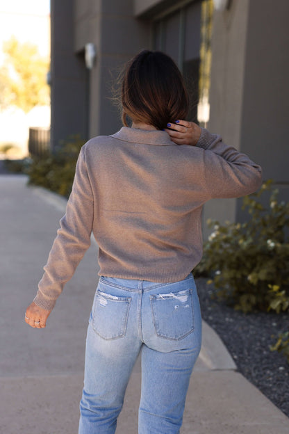 A woman with long brown hair walks outdoors, facing away, wearing a Mocha Button Sweater Up Jacket and light blue jeans.