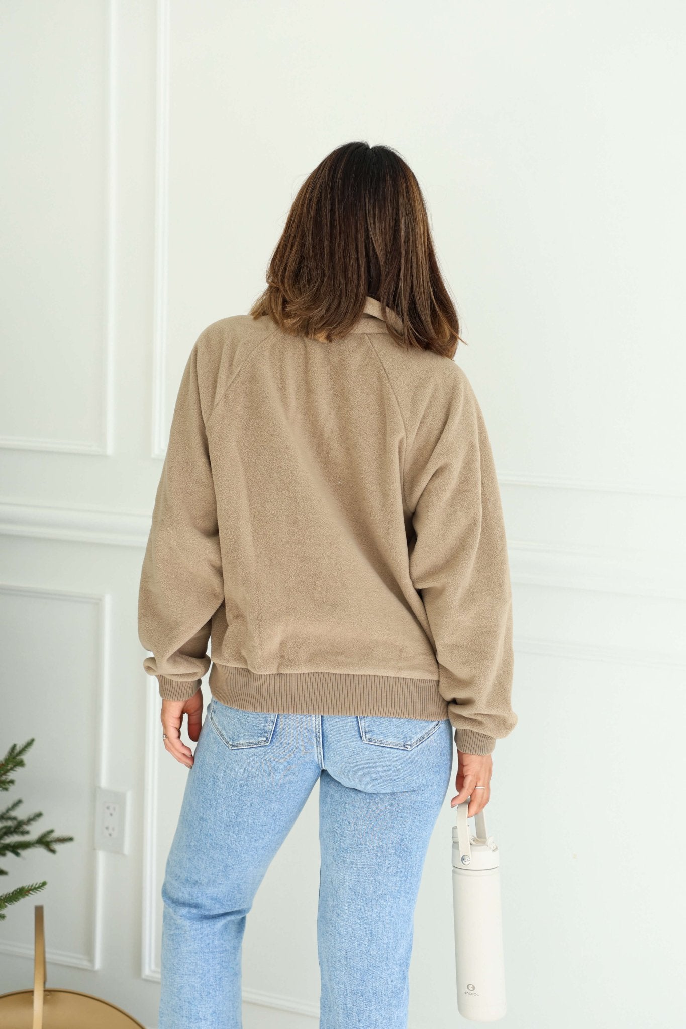 A person with shoulder-length brown hair wears a Mocha Button Up Fleece Pullover and light blue jeans, holding a white bottle.