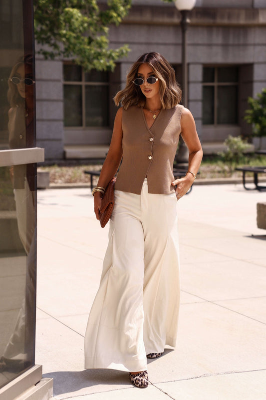 Woman wearing the Mocha Button Up Sweater Vest, white wide-leg pants, clutch, and sunglasses on a sunny day.