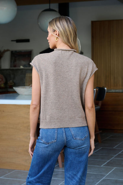 A woman with blonde hair in a Mocha Cap Sleeve Button Down Top and blue jeans stands indoors, facing away from the camera.