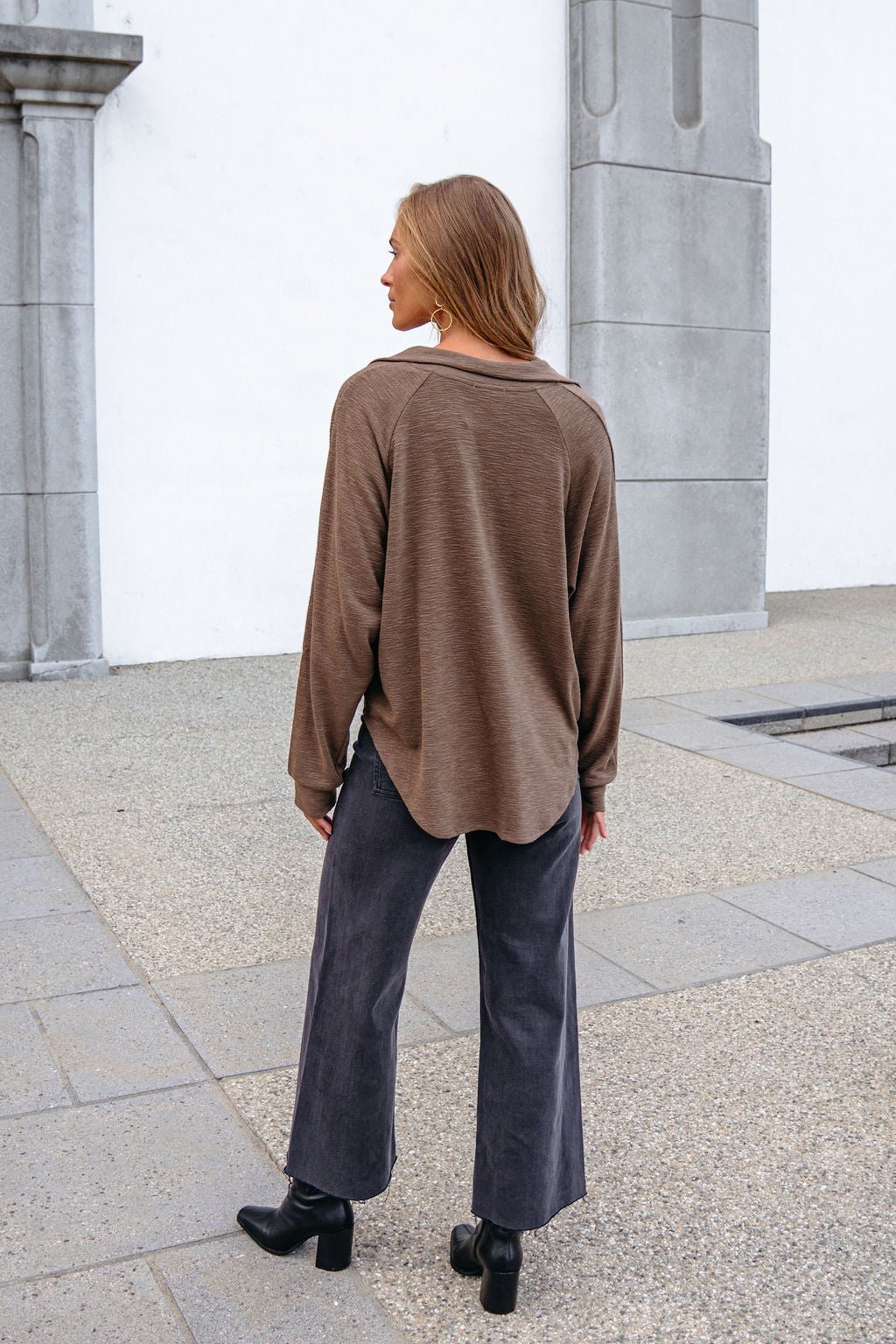 A woman wears a Mocha Collared High Low Pullover, wide-leg black jeans, and boots, standing outdoors with her back to the camera.