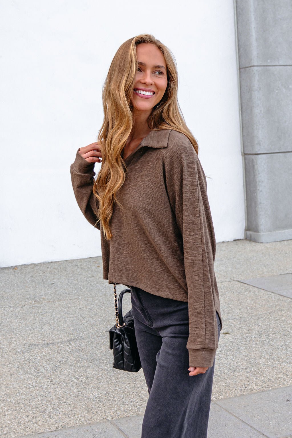 Woman with long hair wears the Mocha Collared High Low Pullover, black pants, and purse, smiling outdoors.