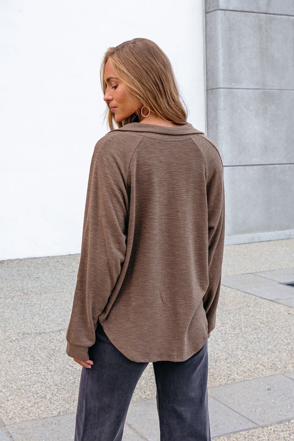 Woman with long hair in a Mocha Collared High Low Pullover and dark pants, outdoors, facing away.