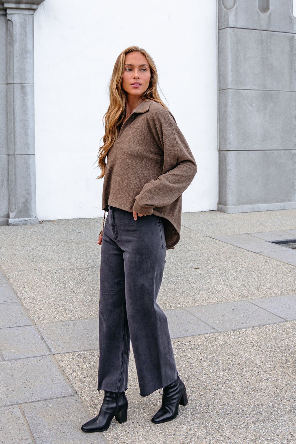 Woman with long hair wears a Mocha Collared High Low Pullover, wide-leg pants, and black heeled boots on the sidewalk.