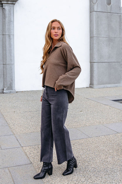 Woman with long hair wears a Mocha Collared High Low Pullover, wide-leg pants, and black heeled boots on the sidewalk.