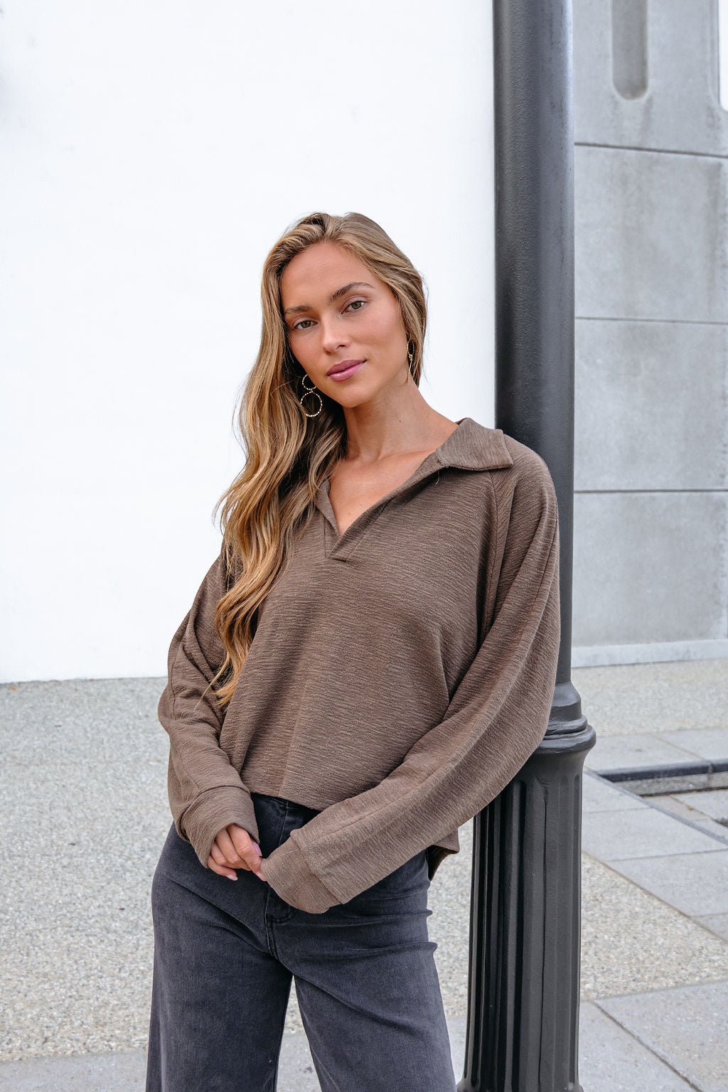 Woman in a Mocha Collared High Low Pullover with an oversized fit poses by a lamppost against a white and gray outdoor wall.