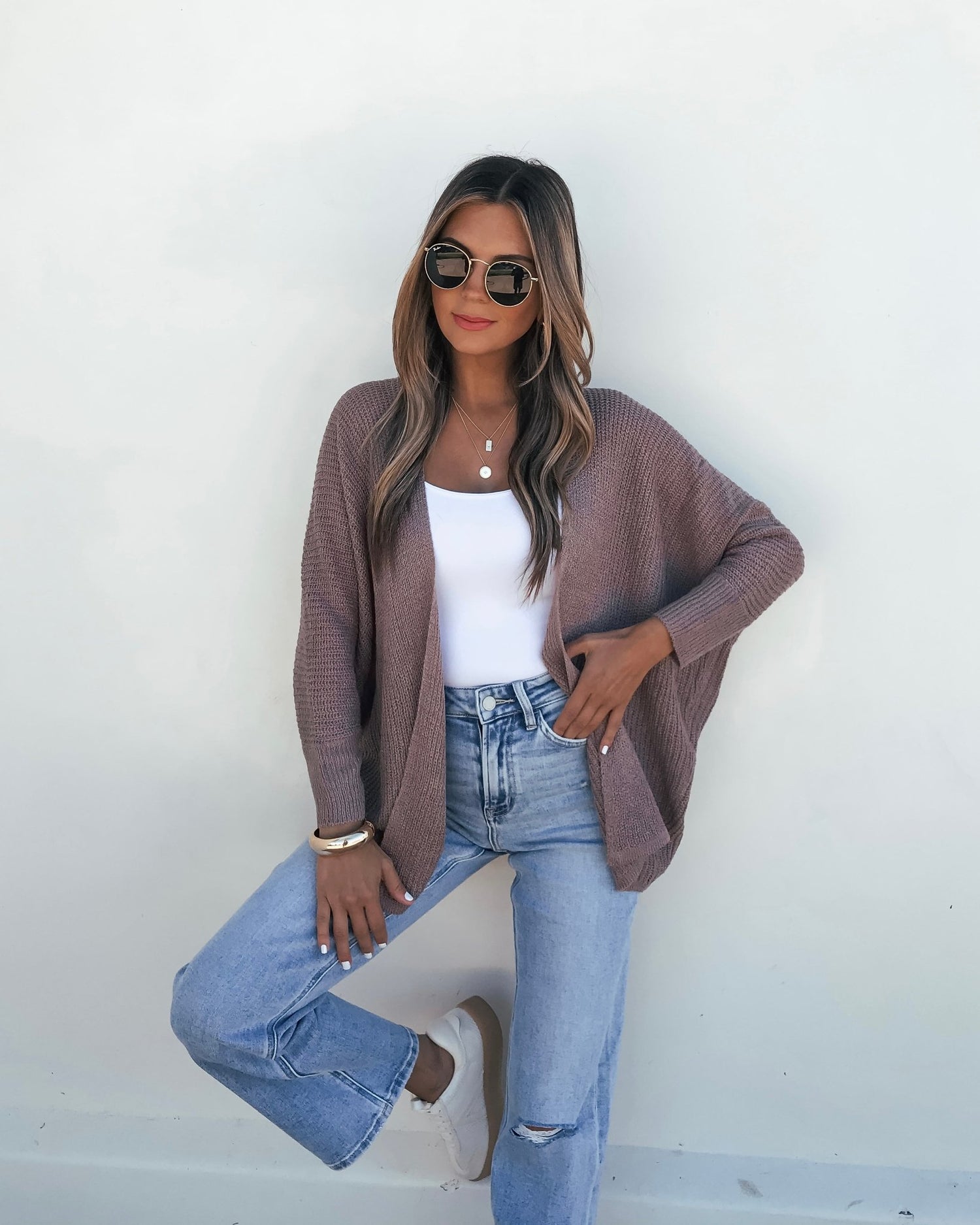 Woman in sunglasses and white top models the Mocha Dolman Sleeve Open Front Cardigan with ripped jeans against a plain white wall.