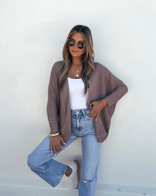 Woman in sunglasses and white top models the Mocha Dolman Sleeve Open Front Cardigan with ripped jeans against a plain white wall.