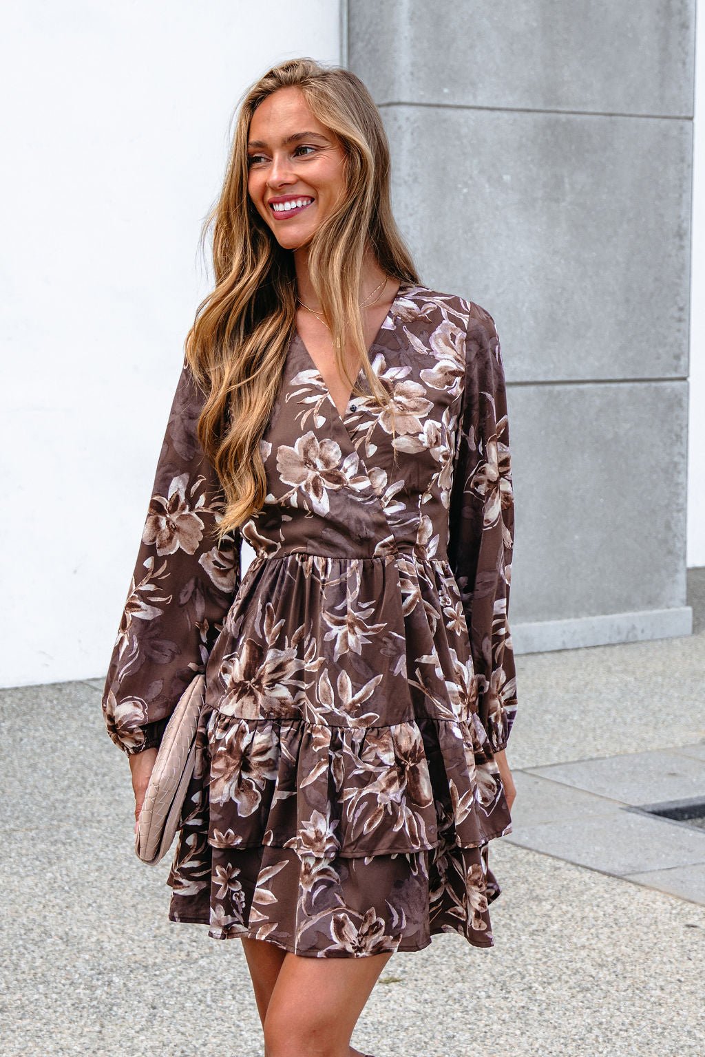 Woman smiling in a Mocha Floral Print Mini Dress with surplice V neckline, holding a beige clutch by a gray wall.