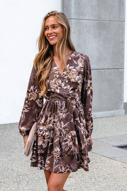Woman smiling in a Mocha Floral Print Mini Dress with surplice V neckline, holding a beige clutch by a gray wall.