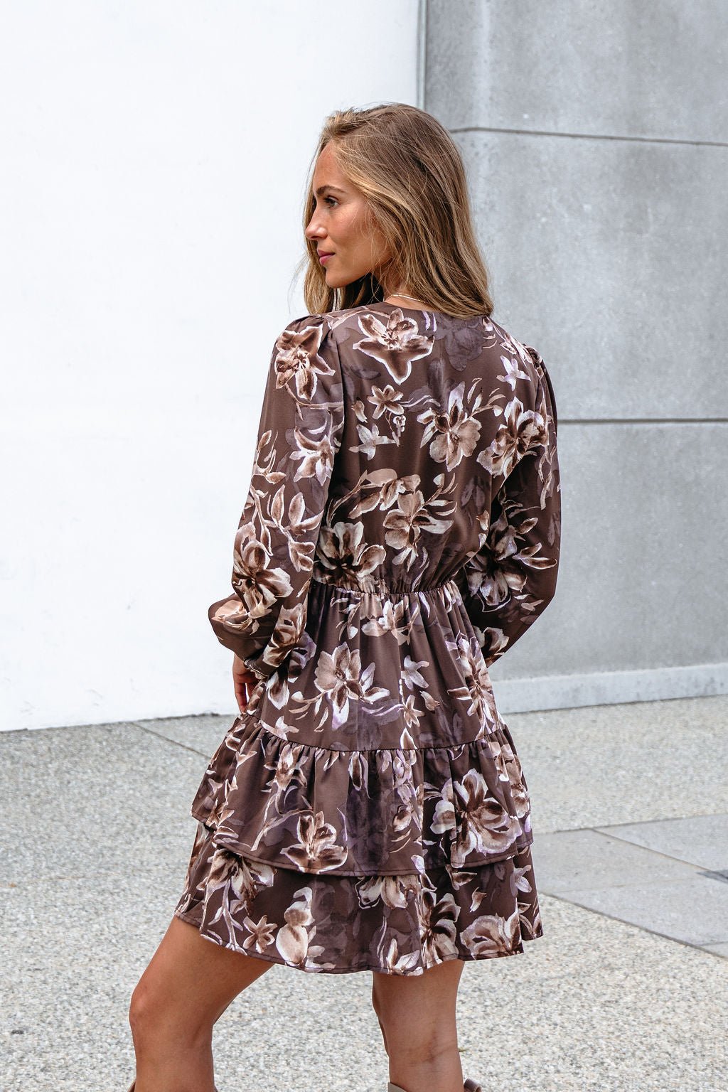 Woman in a Mocha Floral Print Mini Dress with a tiered hem stands outdoors, facing away and turning her head by a concrete wall.