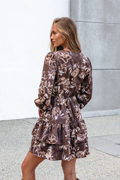 Woman in a Mocha Floral Print Mini Dress with a tiered hem stands outdoors, facing away and turning her head by a concrete wall.