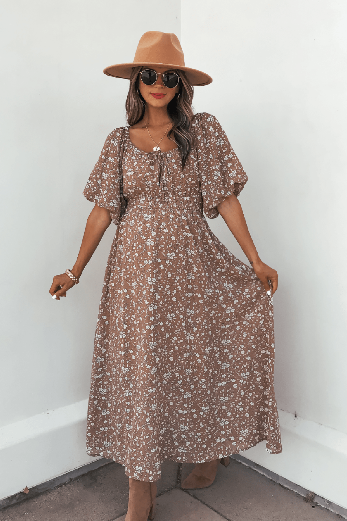 Woman in a tan hat and sunglasses wears the Mocha Floral Print Smocked Midi Dress, standing against a white wall holding out her dress.