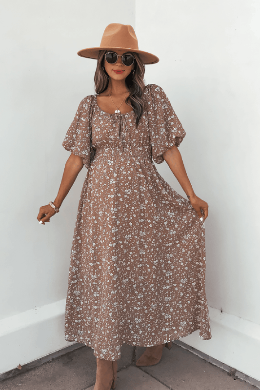 Woman in a tan hat and sunglasses wears the Mocha Floral Print Smocked Midi Dress, standing against a white wall holding out her dress.
