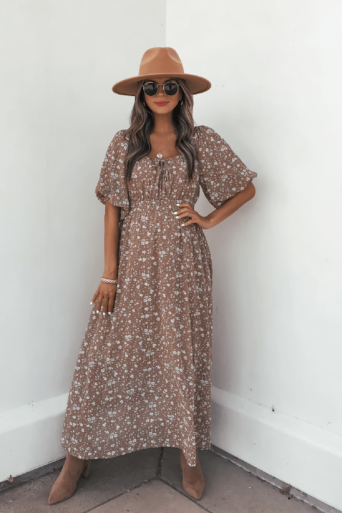 Woman in Mocha Floral Print Smocked Midi Dress, tan hat, and sunglasses poses by a white wall with one hand on her hip.